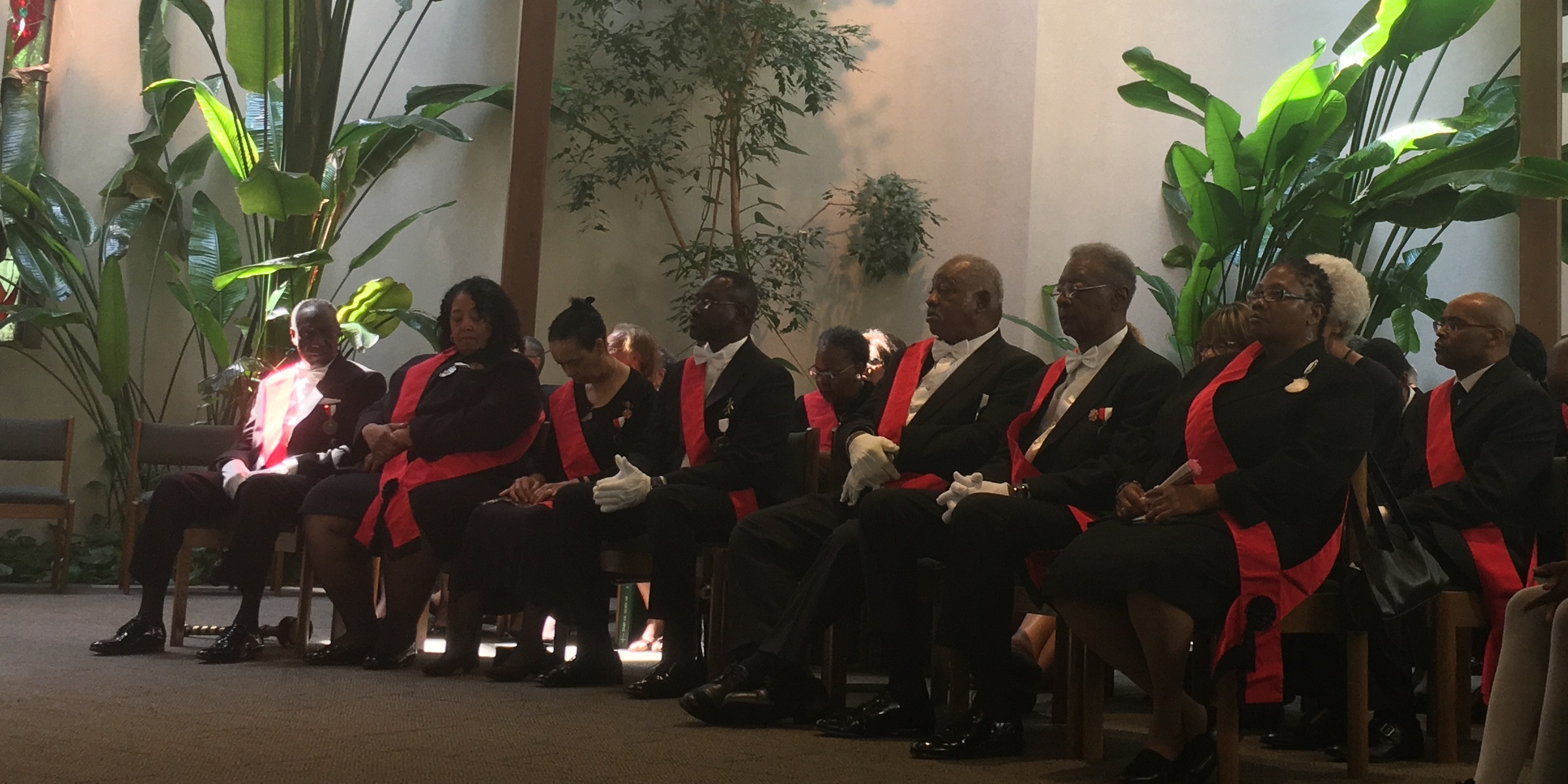 row of seated members of Catholic dressed in black with red sash during Mass