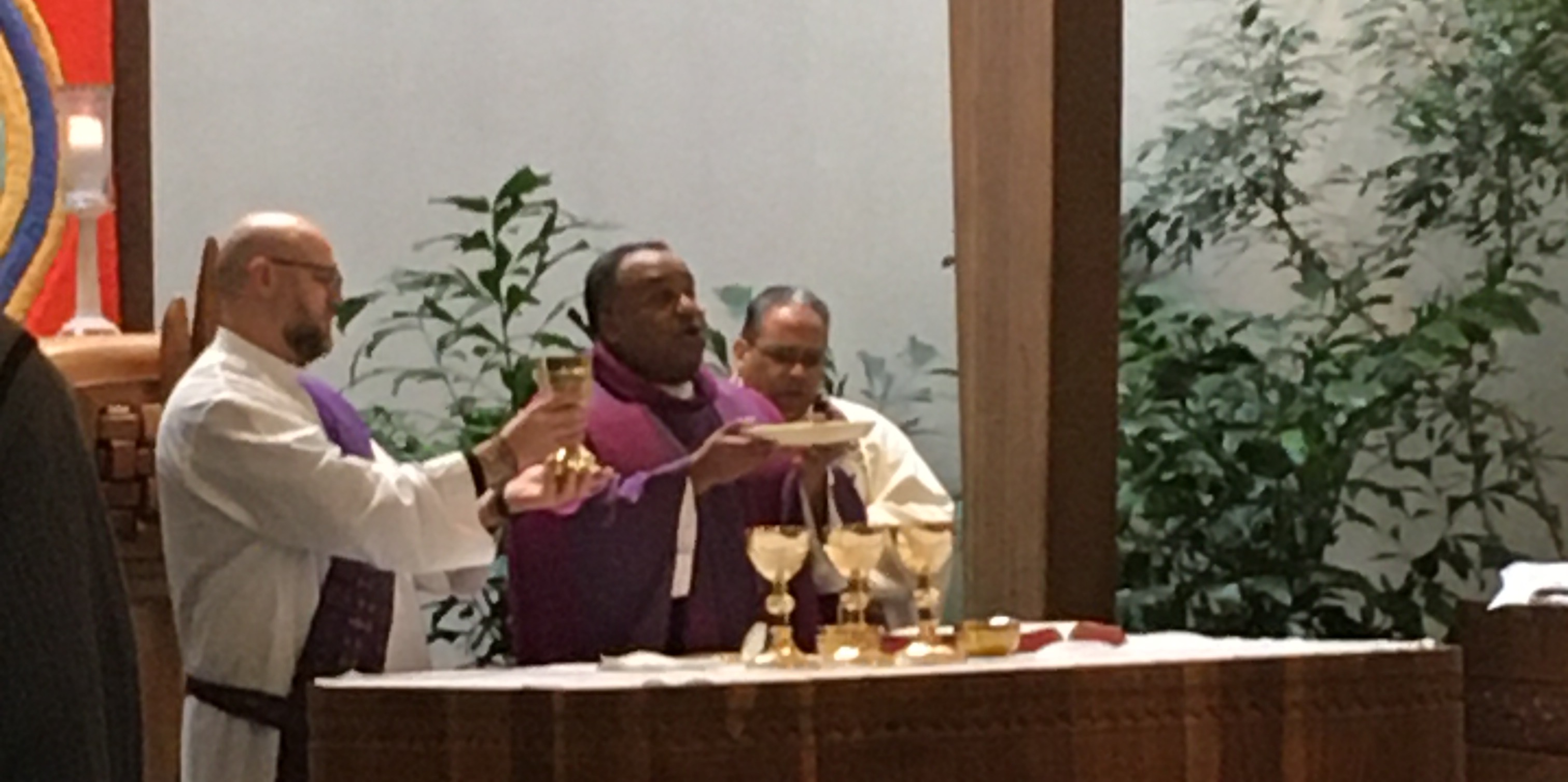 Father Jones with bread and wine in front of Catholic altar with deacon and priest on sides