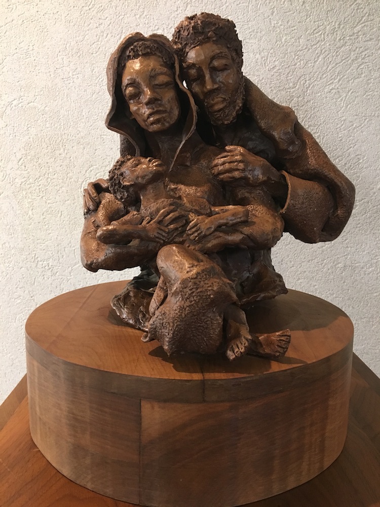 Holy family wood sculpture of Joseph embracing Mary who is embracing the child Jesus.